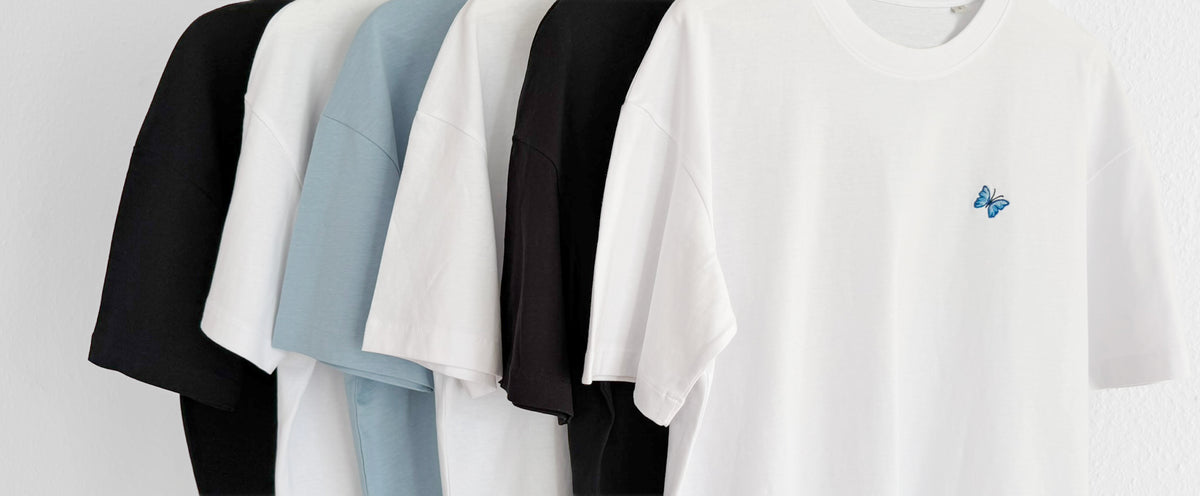 Zu erkennen sind die unterschiedlichen Farben, die im Shop angeboten werden. Im Vordergrund hängt ein weißes Shirt mit gesticktem Schmetterling Motiv