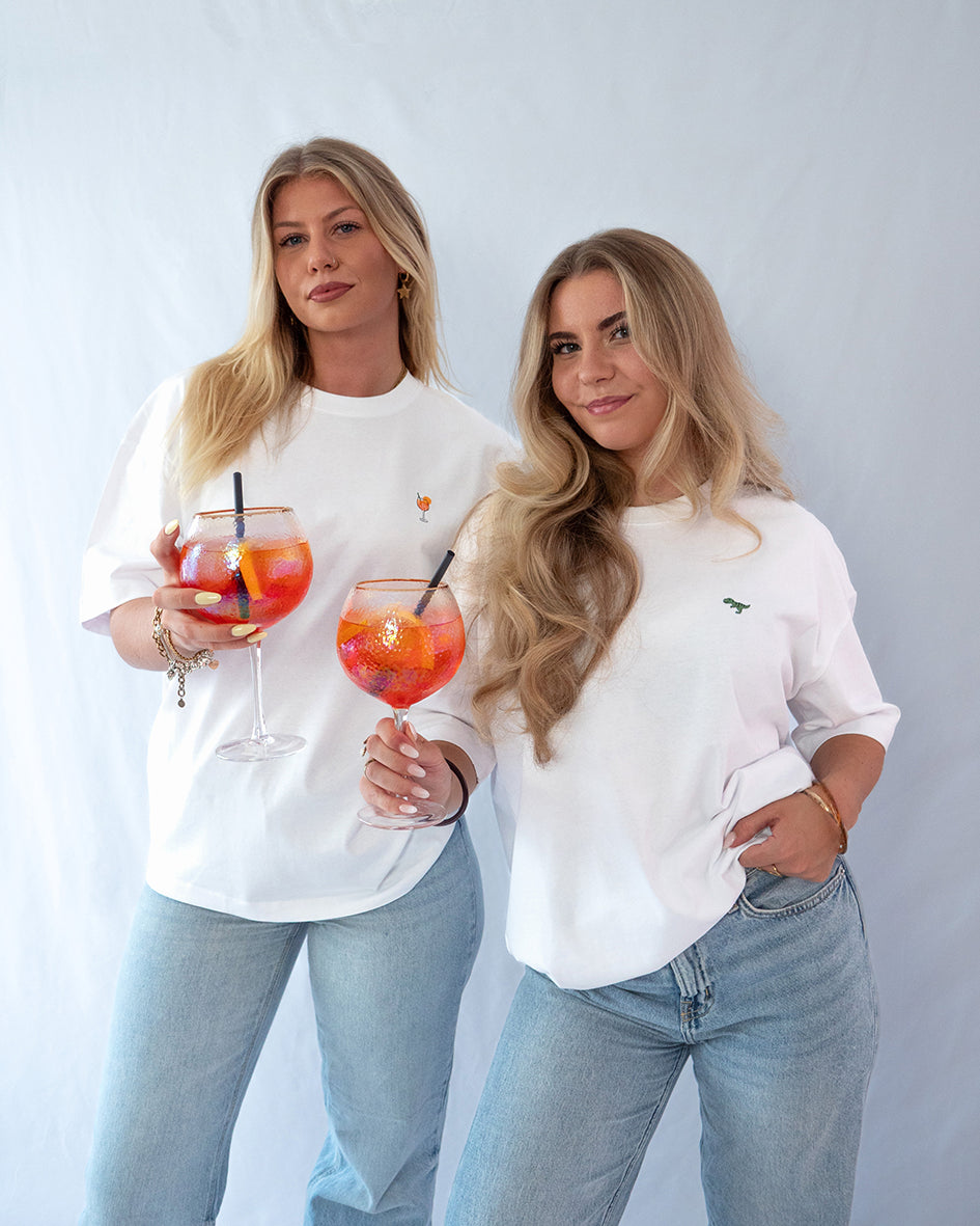 Beide Model tragen ein weißes Bio-Baumwoll Shirt, eine mit gesticktem T-Rex Motiv, die andere mit gesticktem Aperol Spritz Motiv auf der Brust