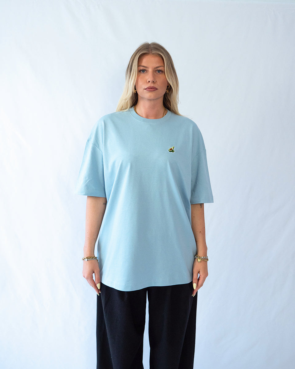 Das Model trägt ein blaues Baumwoll Shirt mit dezent gesticktem Avocado Motiv auf der Brust