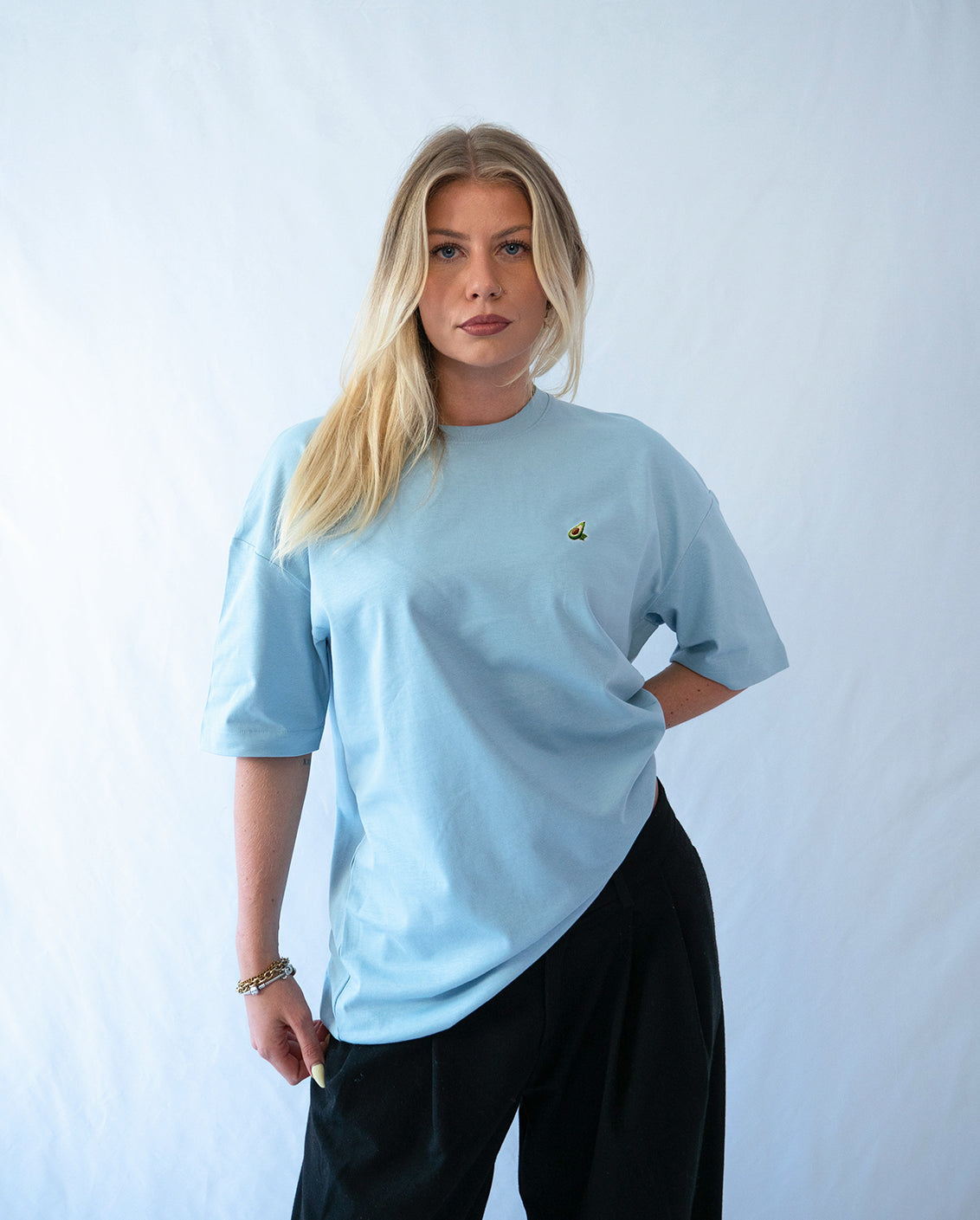 Das Model trägt ein blaues Bio-Baumwoll T-Shirt mit gesticktem Avocado Motiv auf der Brust
