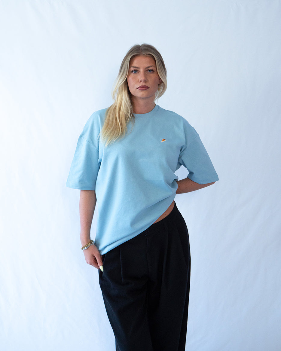 Das Model trägt ein blaues Bio-Baumwoll T-Shirt mit gesticktem Pizza Slice Motiv auf der Brust