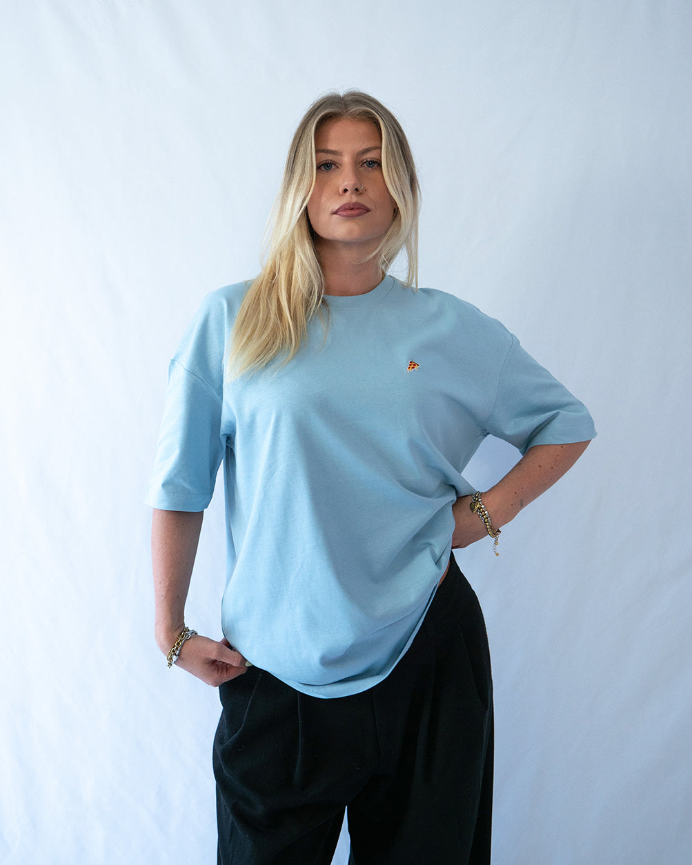 Das Model trägt ein blaues Bio-Baumwoll T-Shirt mit gesticktem Pizza Slice Motiv auf der Brust