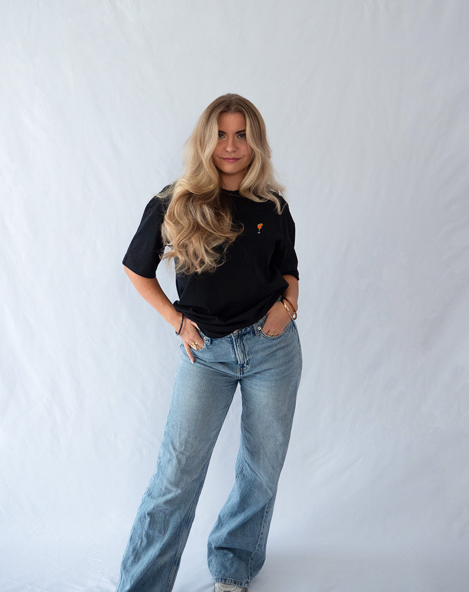 Das Model trägt ein schwarzes Bio-Baumwoll Shirt mit dezent gesticktem Aperol Spritz Motiv auf der Brust