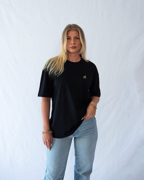 Das Model trägt ein schwarzes Baumwoll Shirt mit gesticktem Avocado Motiv auf der Brust
