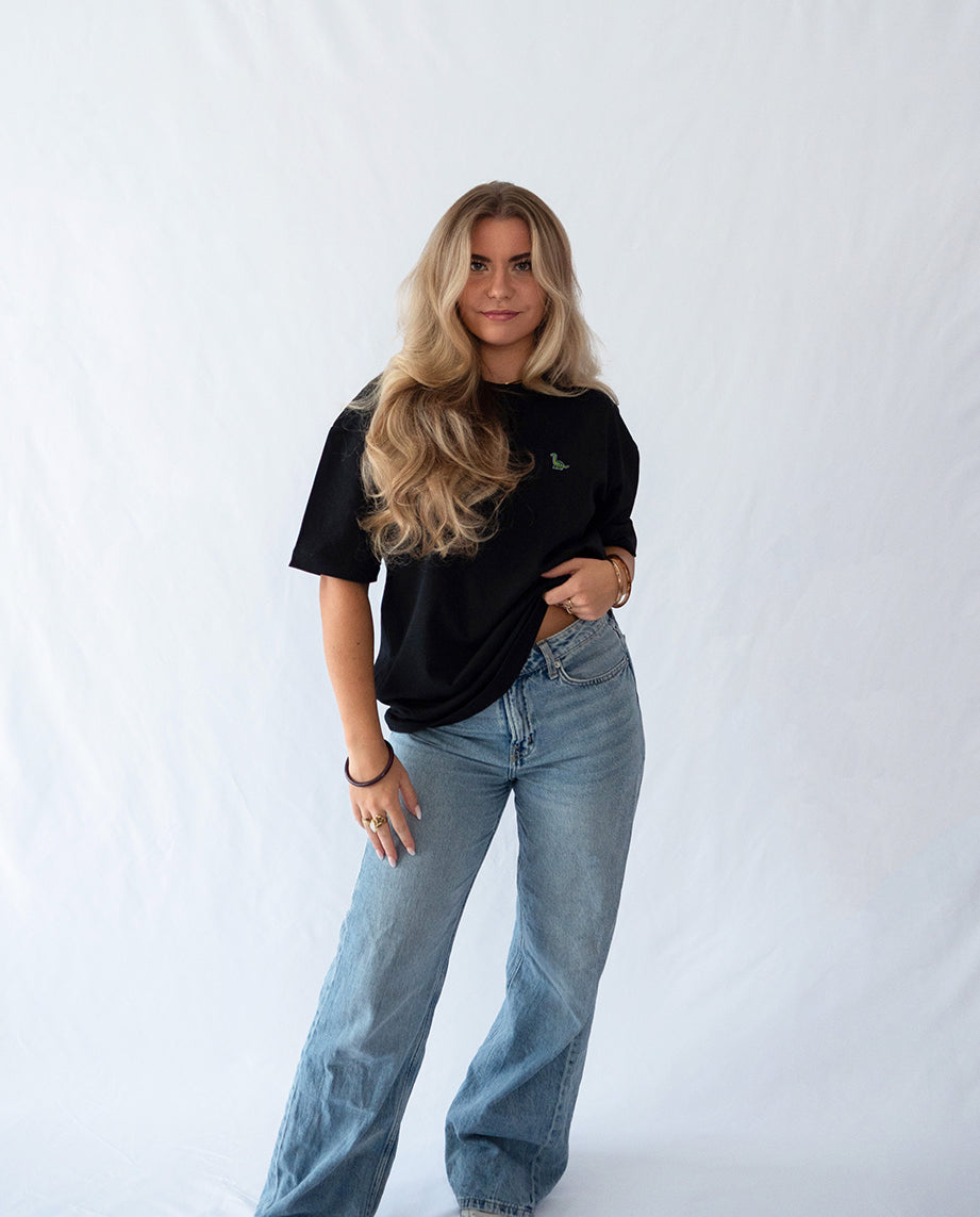 Das Model trägt ein schwarzes oversize Bio-Baumwoll T-Shirt mit gesticktem Brontosaurus Motiv auf der Brust