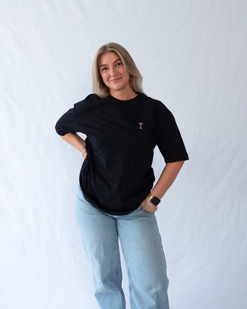 Das Model trägt ein schwarzes Bio-Baumwoll T-Shirt mit gesticktem Espresso Martini Cocktail Motiv auf der Brust