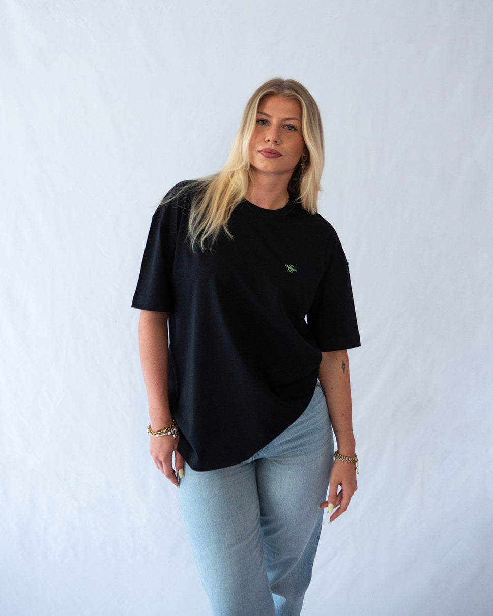 Das Model trägt ein schwarzes oversize Bio-Baumwoll T-Shirt mit gesticktem Spinosaurus Motiv auf der Brust