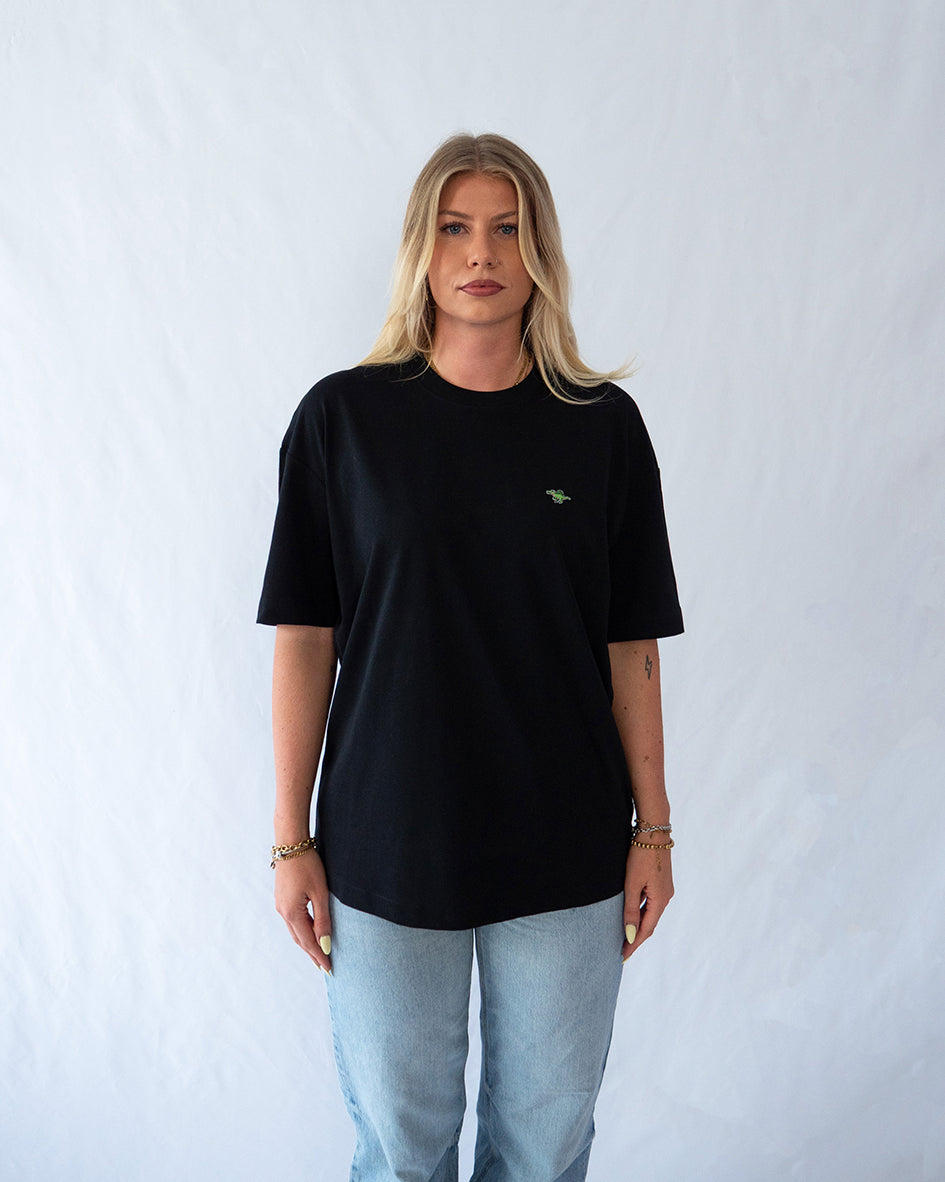 Das Model trägt ein schwarzes oversize Bio-Baumwoll T-Shirt mit gesticktem Spinosaurus Motiv auf der Brust