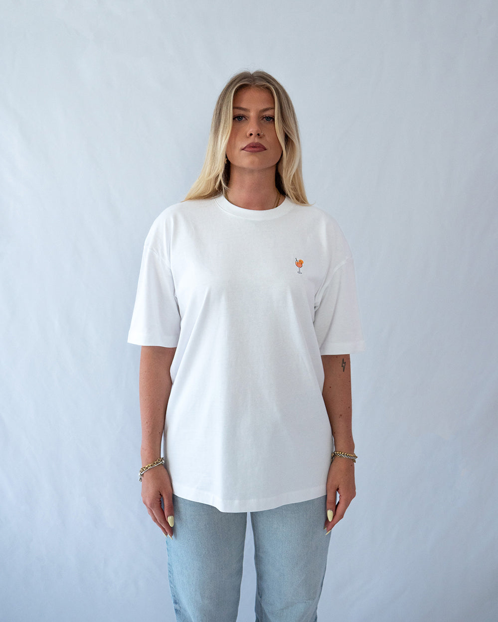Das Model trägt ein weißes Bio-Baumwoll Shirt mit dezent gesticktem Aperol Spritz Motiv auf der Brust