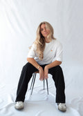 Das Model trägt ein weißes Bio-Baumwoll T-Shirt mit gesticktem Pizza Slice Motiv auf der Brust und sitzt auf einem Hocker