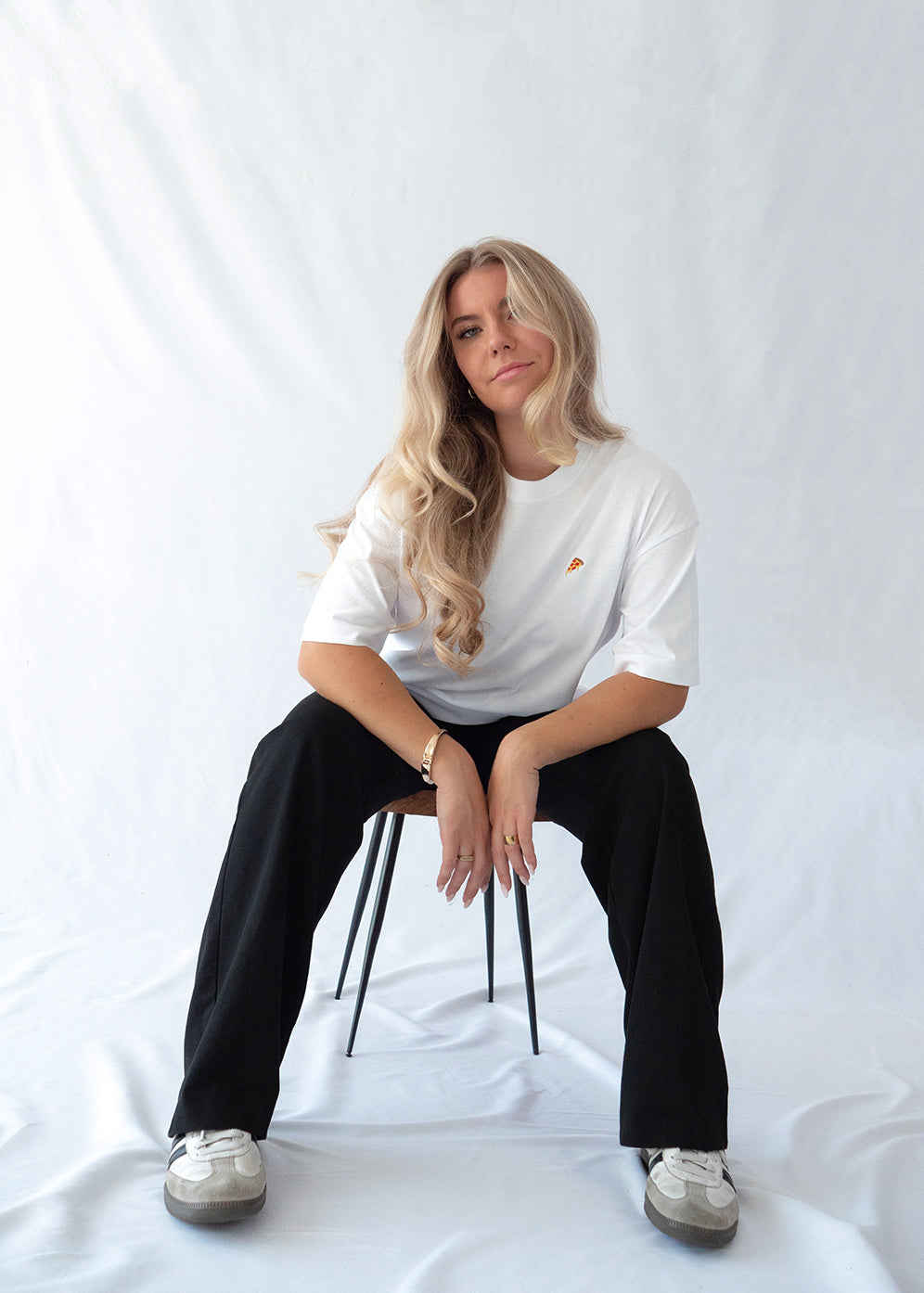 Das Model trägt ein weißes Bio-Baumwoll T-Shirt mit gesticktem Pizza Slice Motiv auf der Brust und sitzt auf einem Hocker