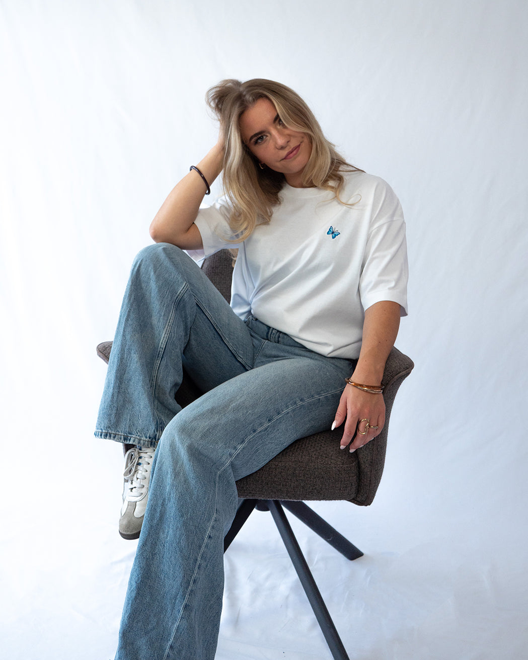 Das Model sitzt auf einem Sessel und trägt ein weißes oversize Baumwoll Shirt mit gesticktem Schmetterling Motiv auf der Brust