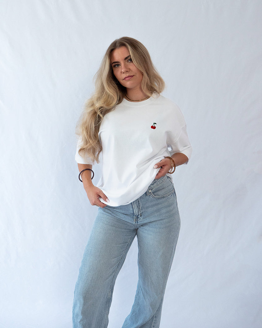 Das Model trägt ein weißes Bio-Baumwoll T-Shirt mit gesticktem Kirschen Motiv auf der Brust