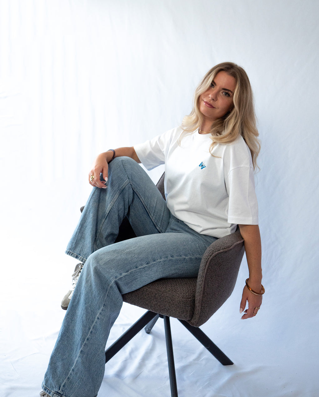 Das Model sitzt auf einem Sessel und trägt ein weißes oversize Baumwoll T-Shirt mit gesticktem Schmetterling Motiv auf der Brust